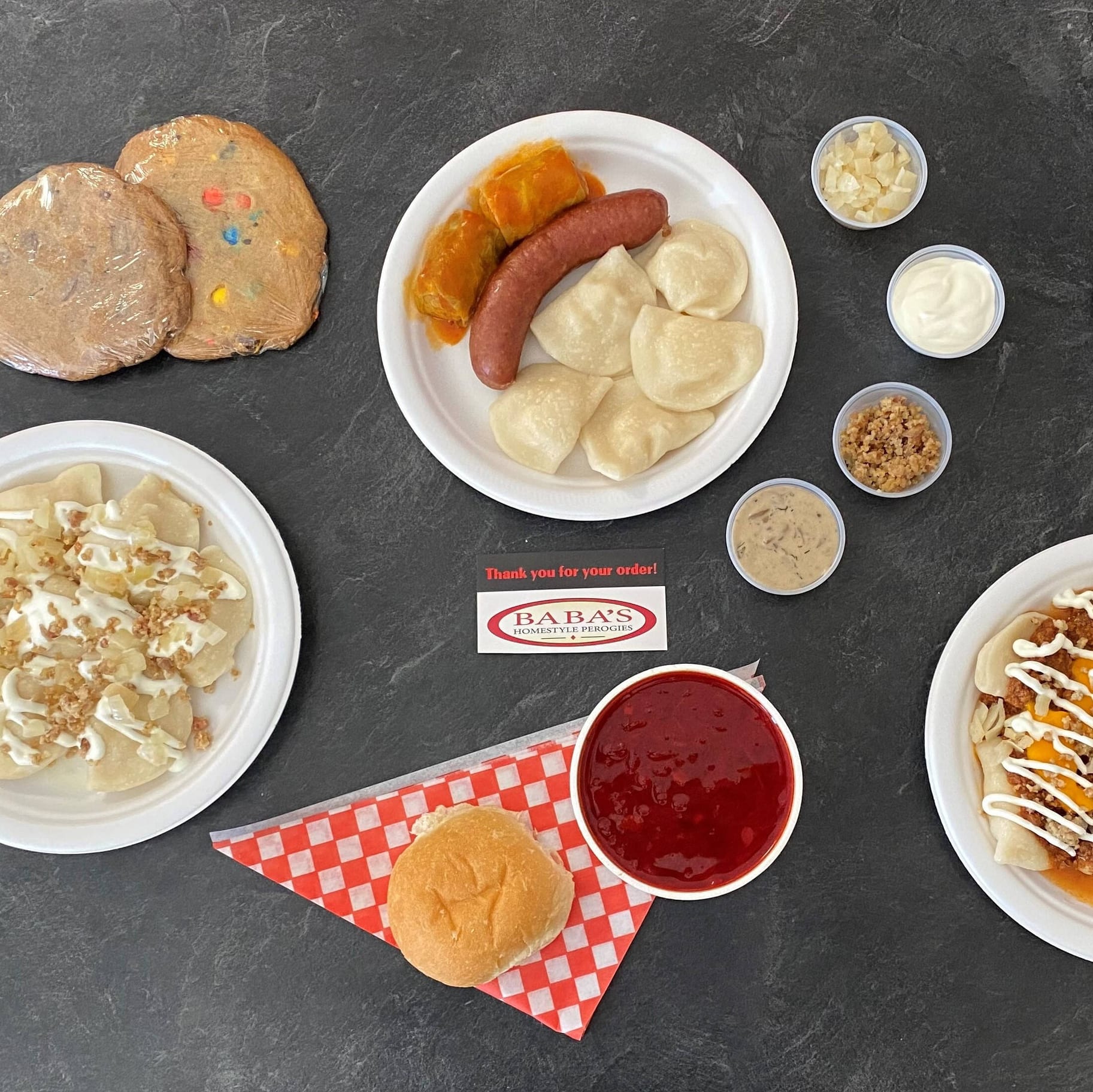 A table of Baba's food from above: plates of perogies and cabbage rolls, a smokie on a bun, borscht, cookies and the fixings